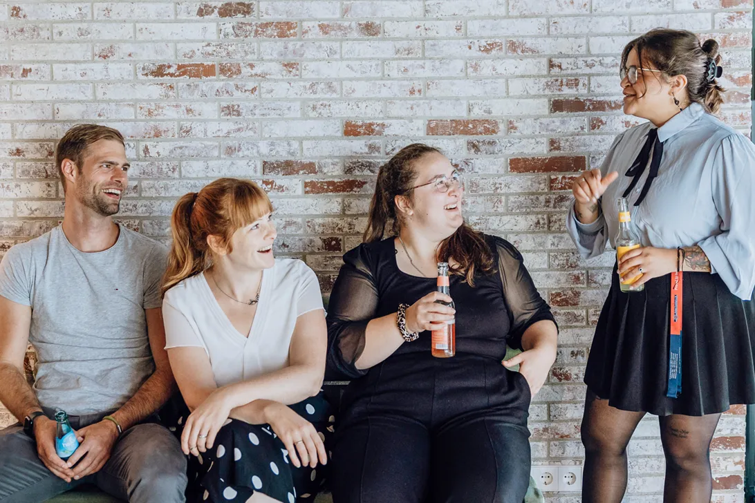 Four people sitting and standing against a brick wall, smiling and holding drinks, engaged in a casual conversation.