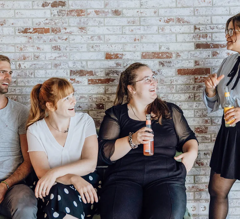 Four people sitting and standing against a brick wall, smiling and holding drinks, engaged in a casual conversation.