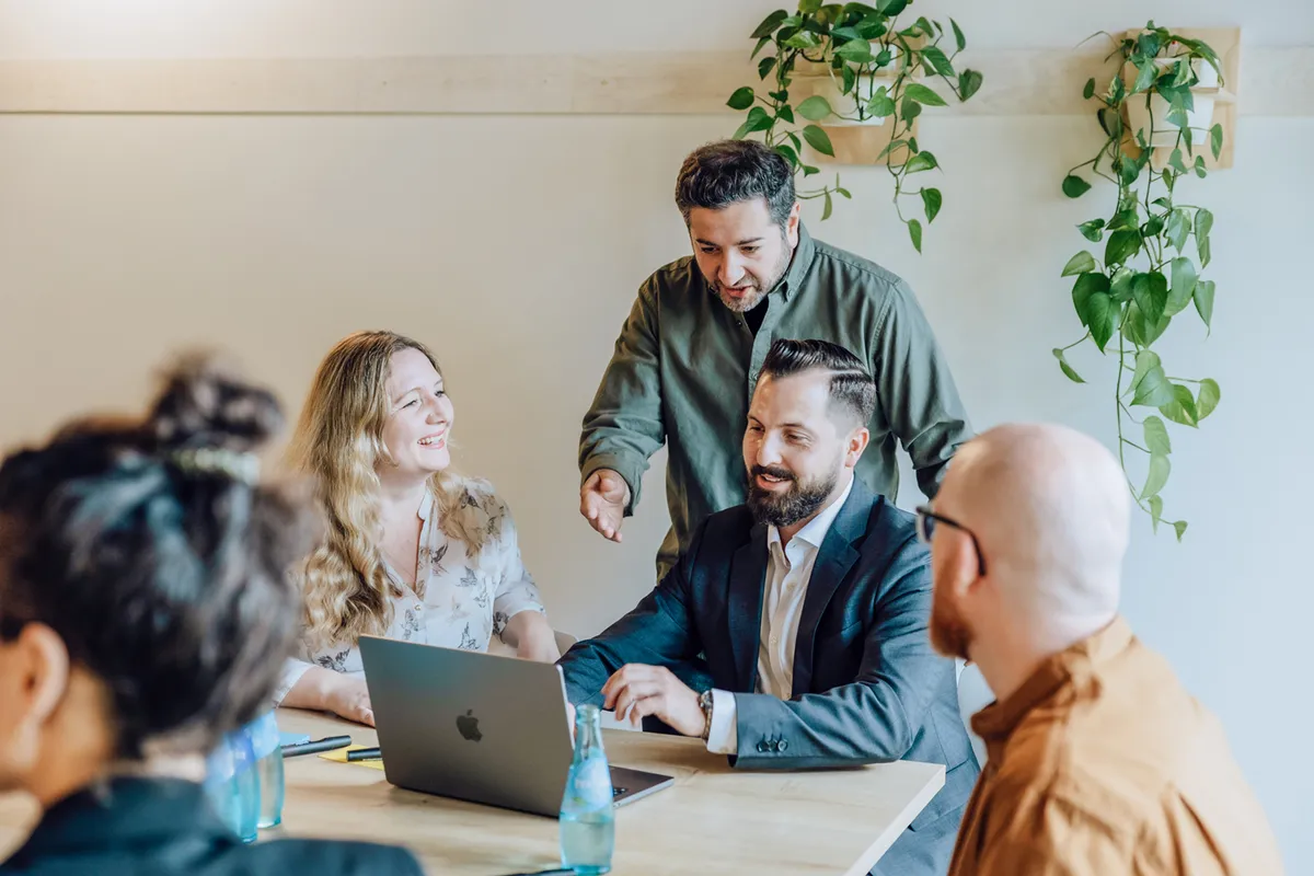 Group of colleagues having a discussion around a table with a laptop and water bottles, surrounded by hanging plants.