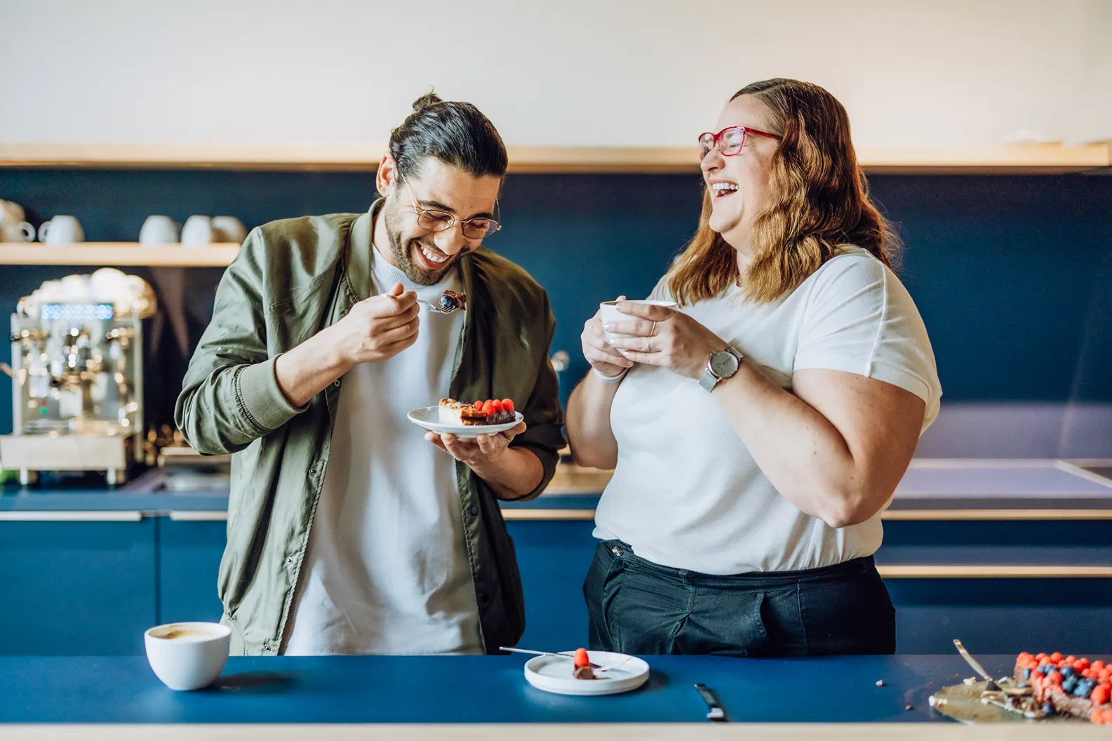 Two people laughing and enjoying dessert in a kitchen setting, with a coffee machine in the background.