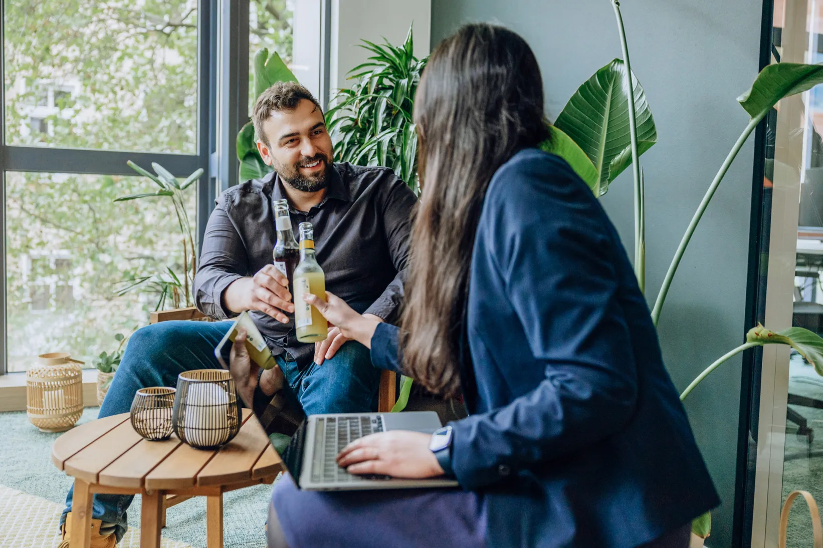 Two people sitting in an office setting with plants, clinking bottles and smiling, one using a laptop.