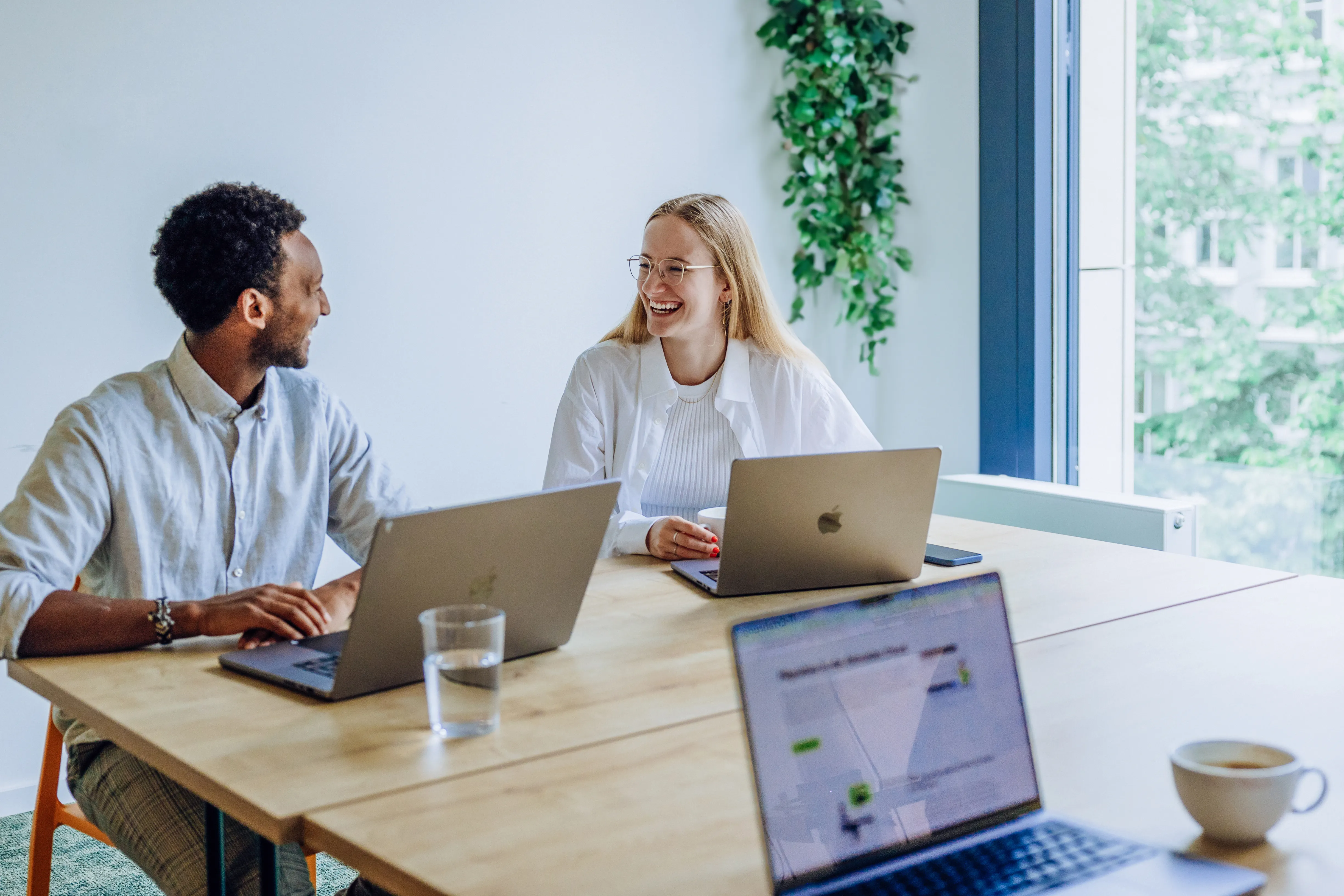 Two people sitting at a table with laptops, smiling and conversing in a bright office space with a window view.