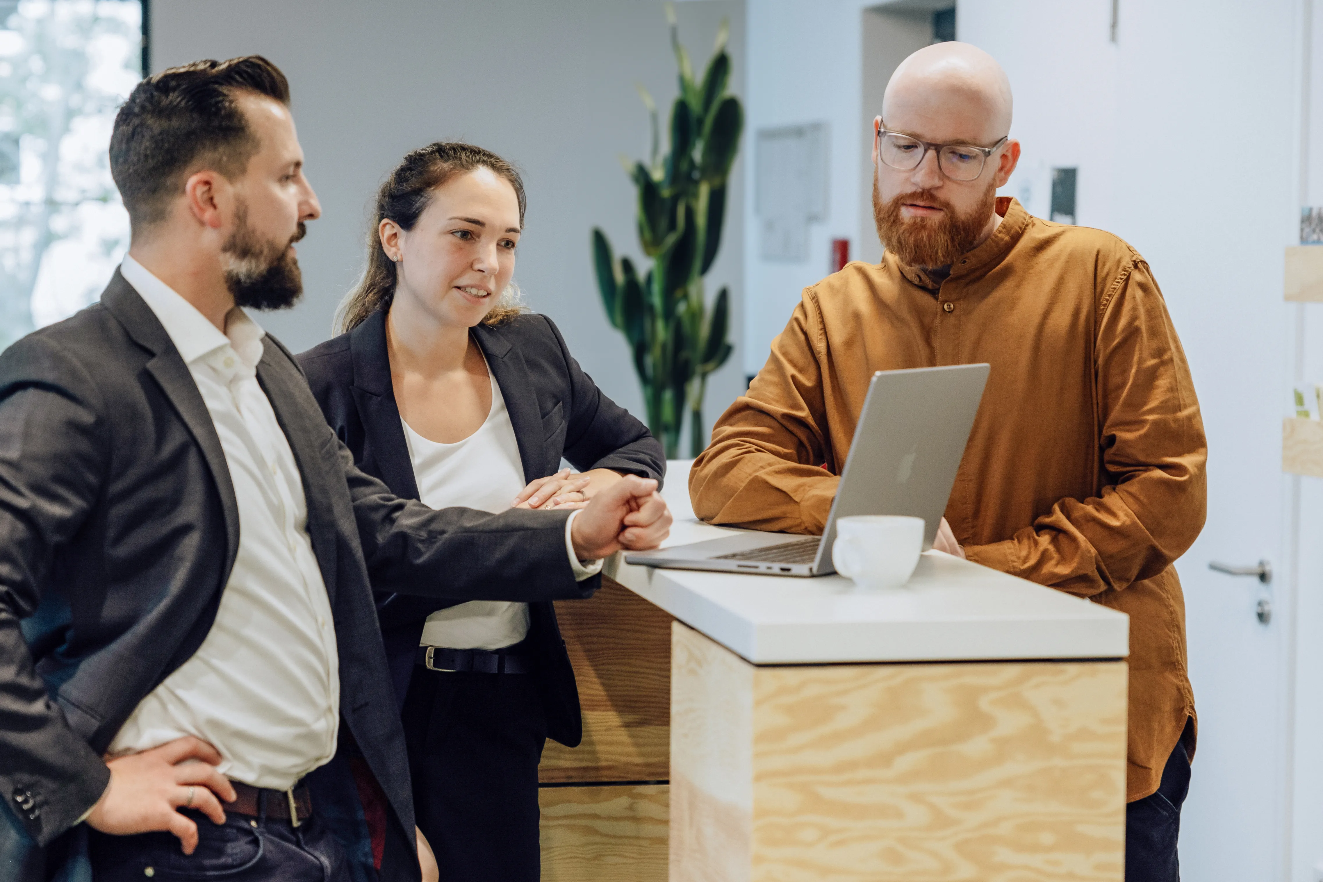 Three people in business attire having a discussion around a laptop on a wooden counter in an office setting.