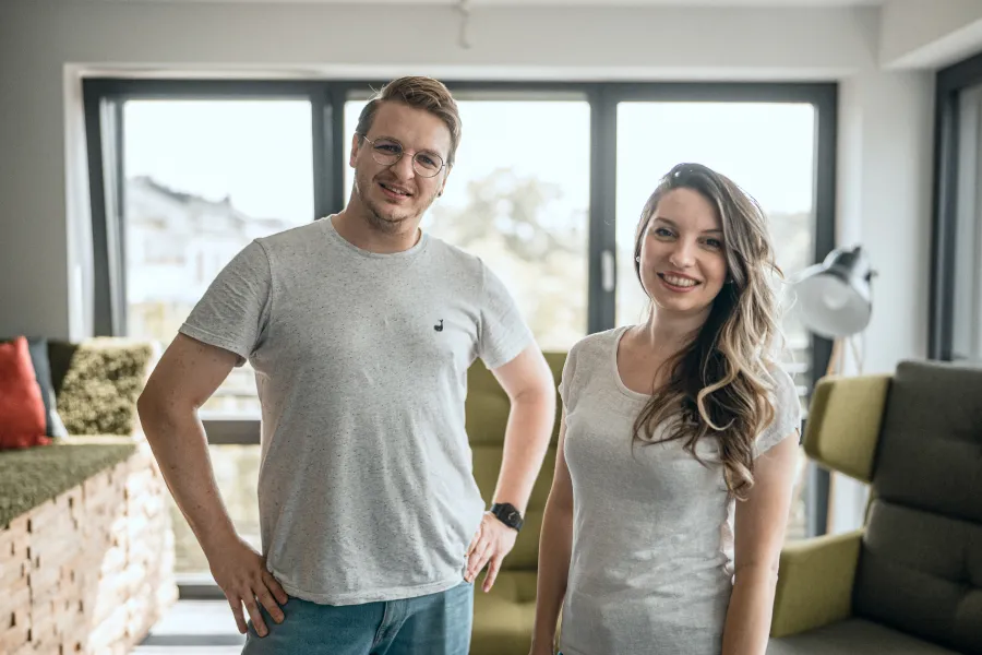 A smiling man and woman wearing casual grey t-shirts stand in a bright office space featuring large windows and green lounge seating in the background.