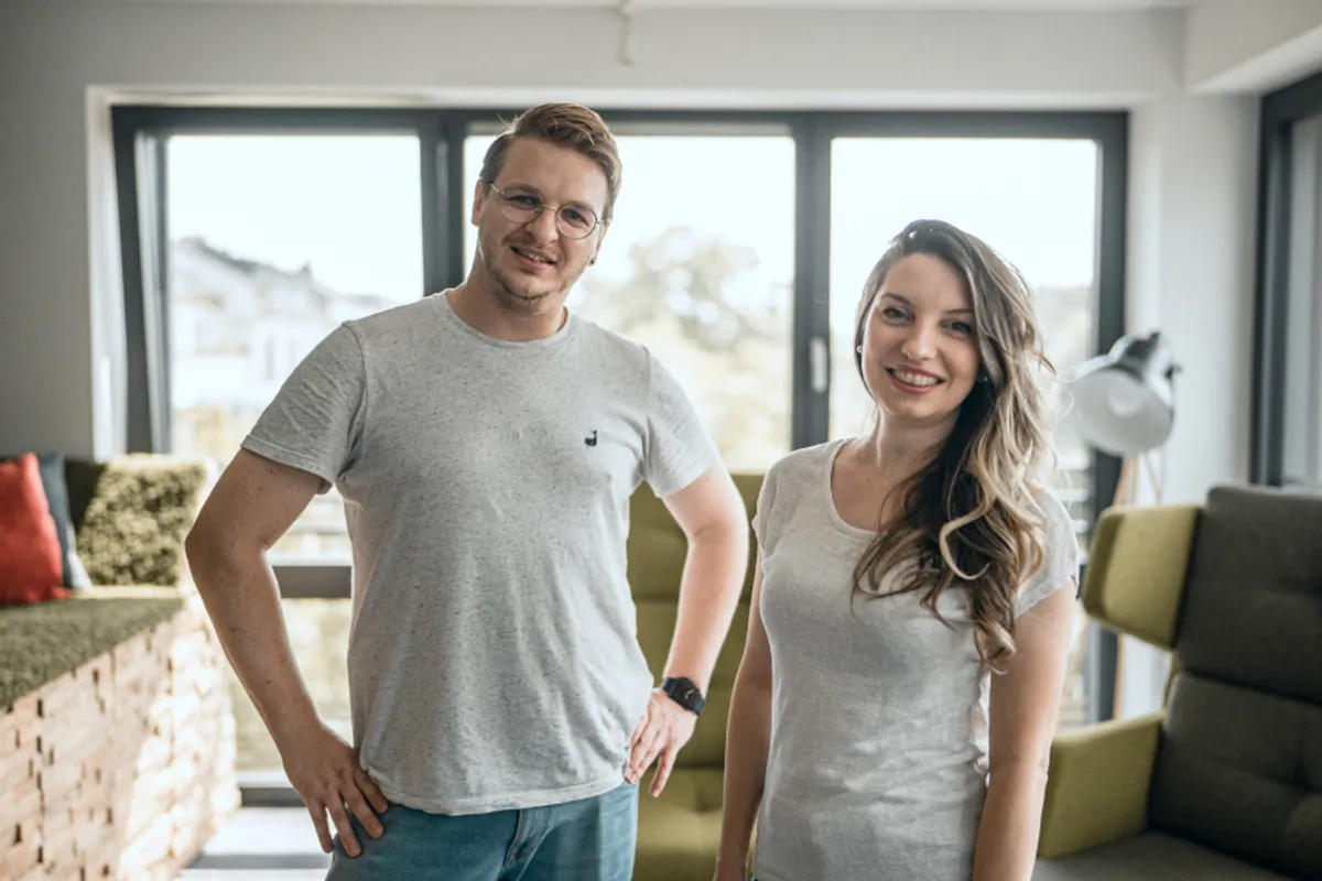 A smiling man and woman wearing casual grey t-shirts stand in a bright office space featuring large windows and green lounge seating in the background.