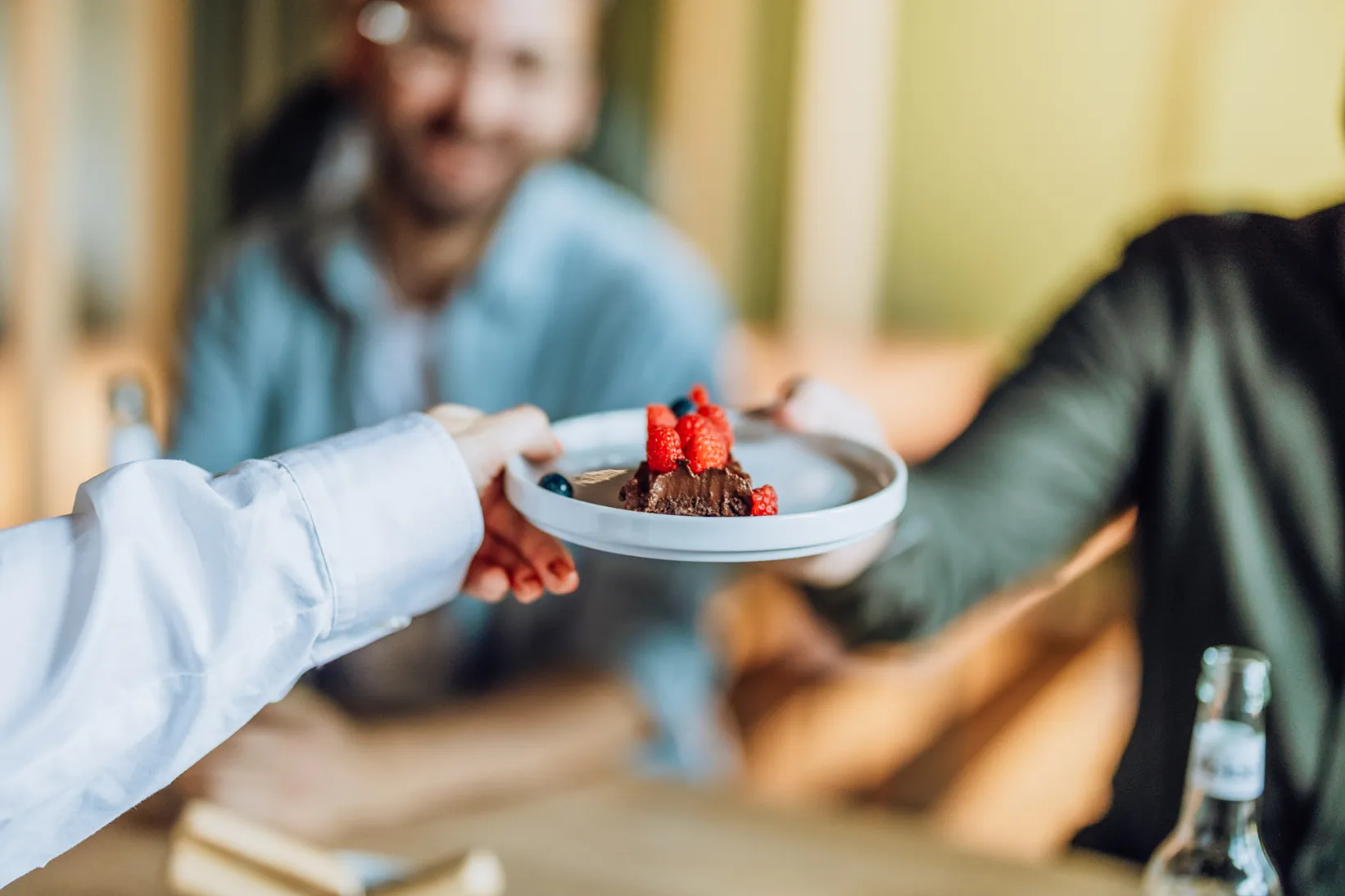 A person handing a plate with a slice of chocolate cake topped with raspberries and blueberries to another person. A blurred figure is smiling in the background.