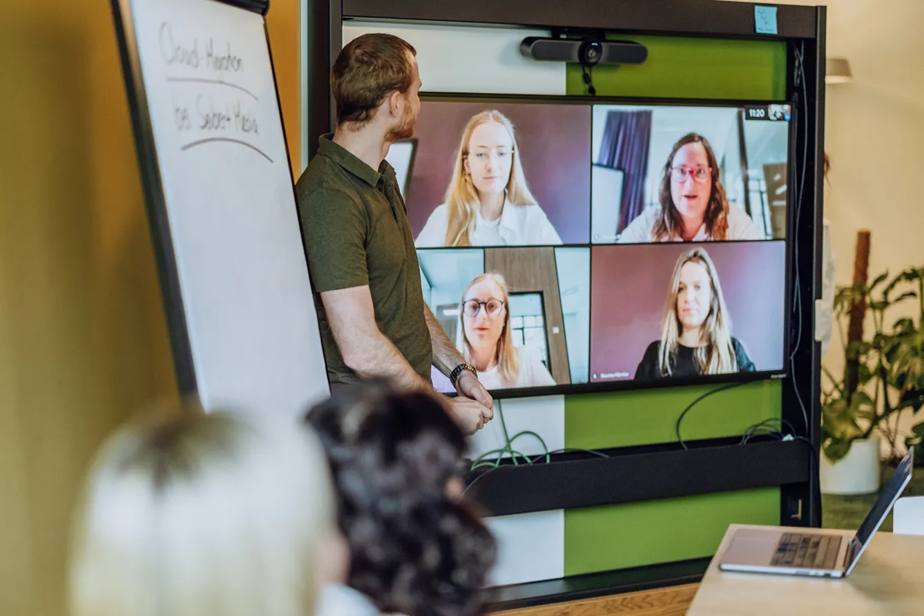 A man standing in front of a large screen displaying a video conference with four participants. A flipchart is visible on the left side of the image.